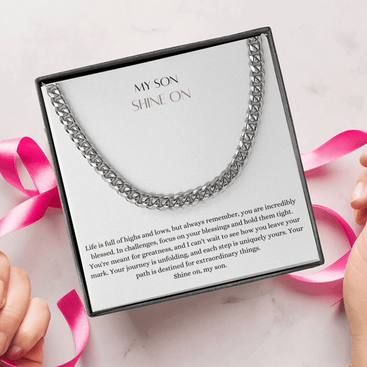 A person unwrapping a necklace gift, with a big Cuban chain necklace with a white gold finish, and a message card that says to my son.