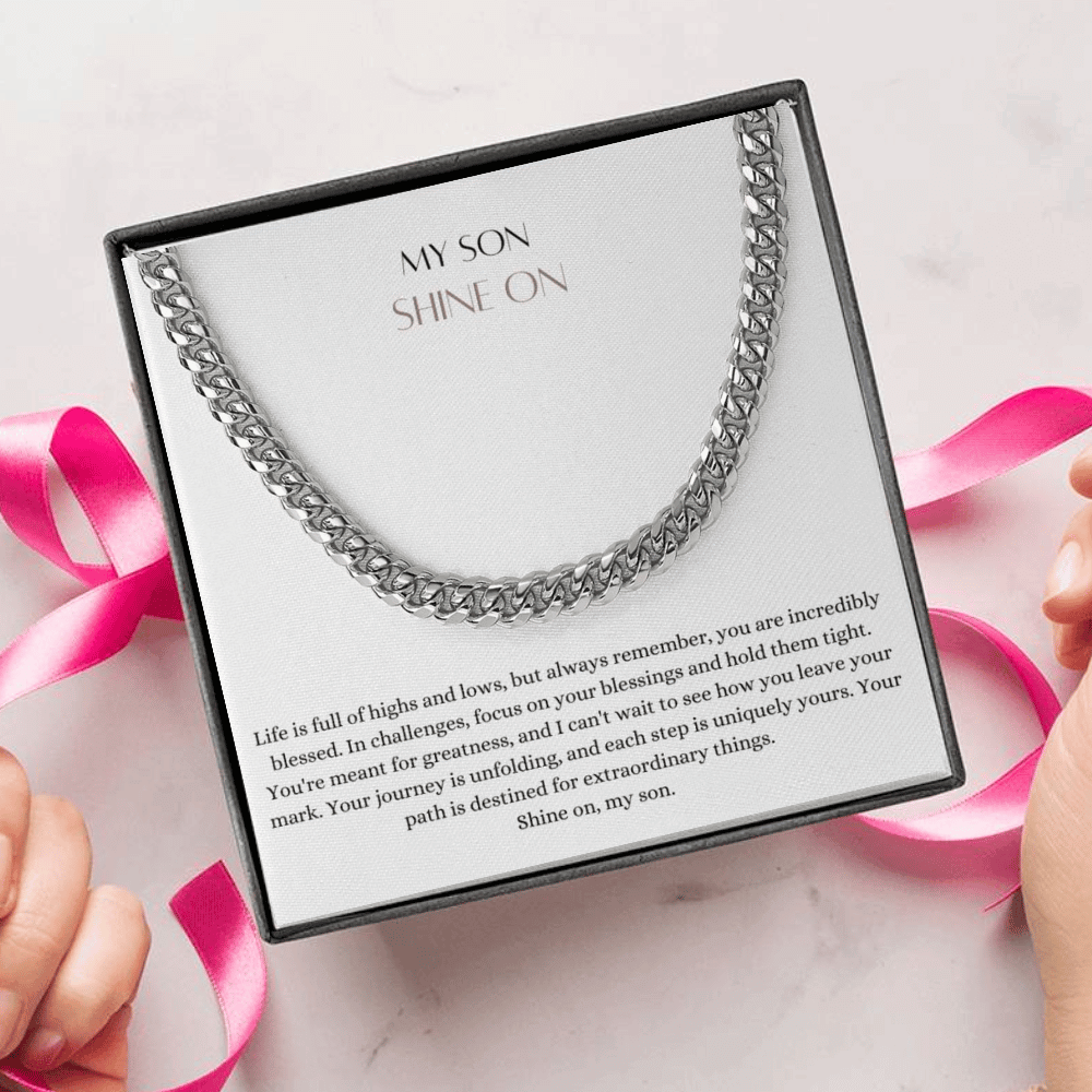 A person unwrapping a necklace gift, with a big Cuban chain necklace with a white gold finish, and a message card that says to my son.