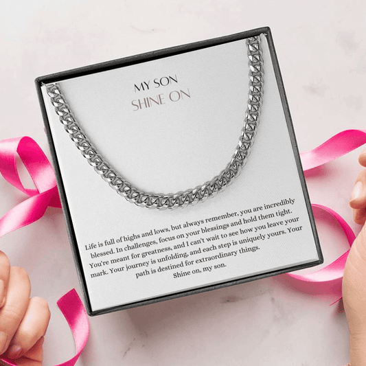 A person unwrapping a necklace gift, with a big Cuban chain necklace with a white gold finish, and a message card that says to my son.