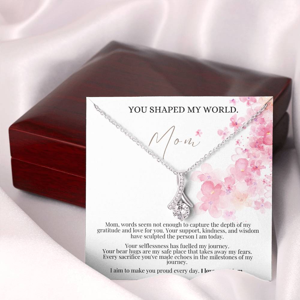 A necklace gift with ribbon shaped pendant with cubic zirconia crystals and white gold finish, with a Mother’s Day message card next to a mahogany gift box.